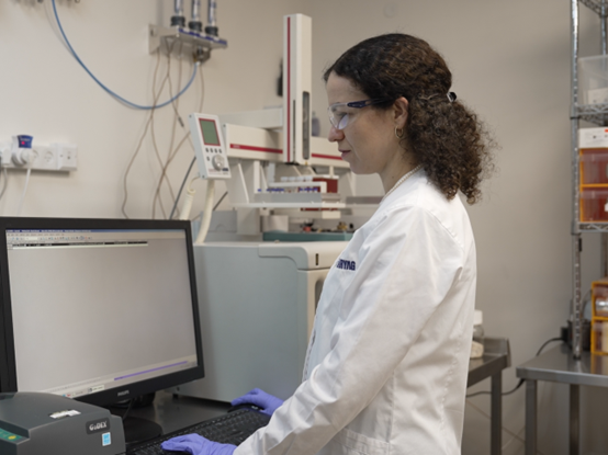 אשת מקצוע במעבדה מבצעת בדיקות אנליטיות באמצעות ציוד מתקדם ומחשב, לבושה חלוק לבן, כפפות ומשקפי מגן בסביבת עבודה סטרילית.